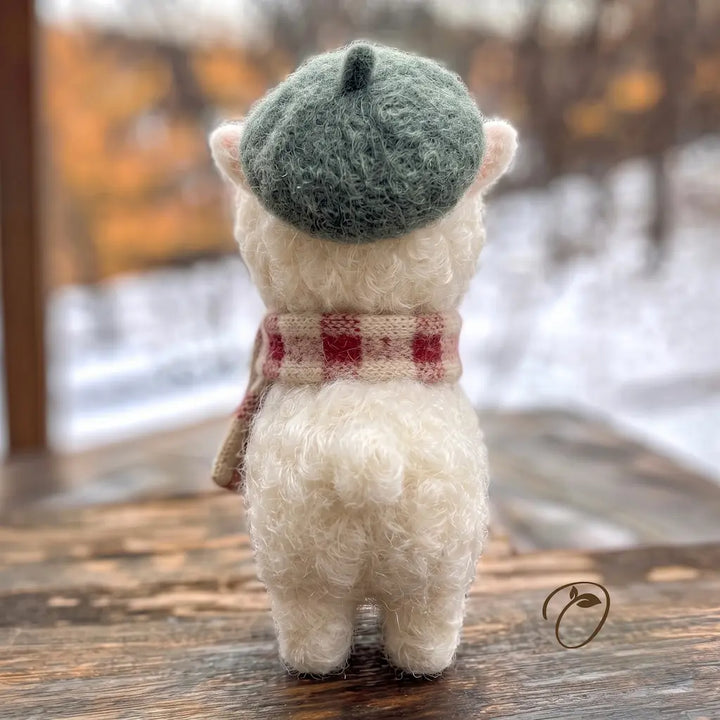 A small white felted wool plush toy shaped like a lamb or goat, shown from the back on a wooden surface. The toy wears a muted green beret and a red-and-beige plaid scarf, with a blurred outdoor winter scene in the background.