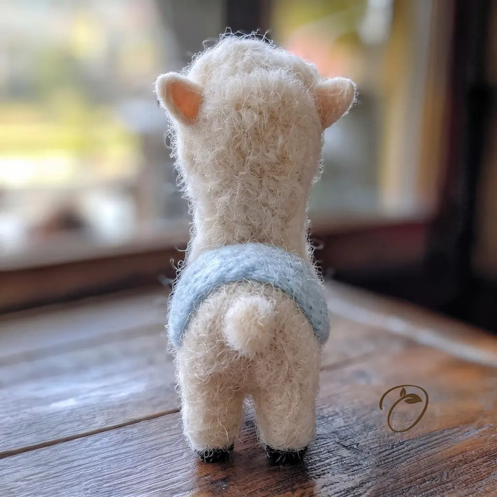 A small fluffy cream-colored felted sheep or alpaca figurine shown from the back, wearing a light blue knitted vest, standing on a wooden surface with a softly blurred indoor background.