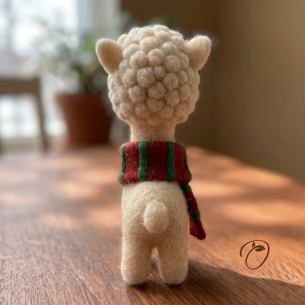 A small felt alpaca toy shown from the back, standing on a wooden table in a softly blurred indoor setting. The alpaca is cream-colored with a red and green striped scarf.