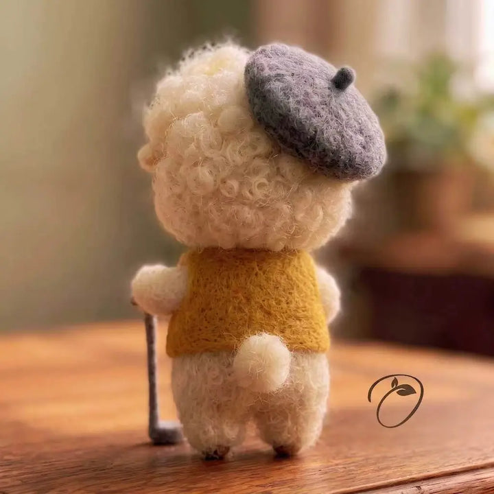 A small wool-felted animal figure shown from the back, standing on a wooden surface. The figure has curly cream-colored wool, a dark gray beret, a mustard yellow top, and holds a thin black cane.