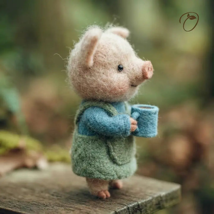 Felted animal figurine of a pig wearing a green vest and blue shirt, holding a cup, with a natural background.