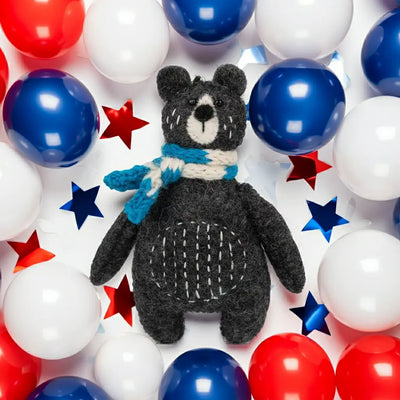 Small black bear plush toy with a blue scarf surrounded by red, white, and blue balloons and stars.