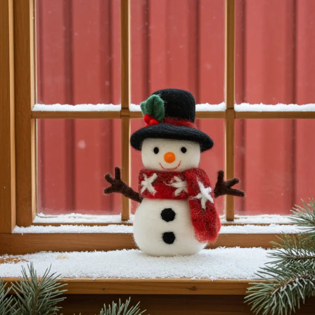Small snowman figurine with a black hat, red scarf, and brown arms, standing on a snowy windowsill in front of a red barn