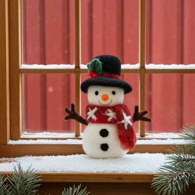 Small snowman figurine with a black hat, red scarf, and brown arms, standing on a snowy windowsill in front of a red barn
