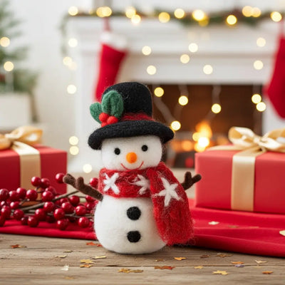 Felted snowman figurine with black top hat and red scarf, sitting on a rustic table in front of a cozy fireplace and red Christmas gifts
