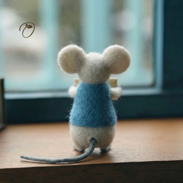 Felted mouse toy wearing a blue sweater on a wooden surface with a blurred window background