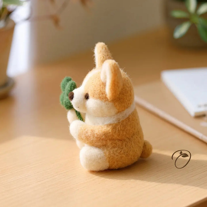 Side view of a seated felt corgi with upright ears, cream paws and a green clover held close to its chest