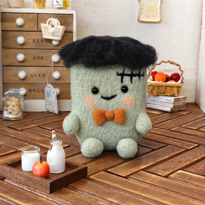 Front view of a sage-green needle-felted Halloween monster with black fuzzy hair, stitched forehead, rosy cheeks and orange bow tie, sitting beside miniature milk bottles and an apple.