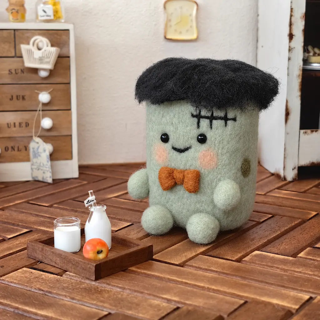 Three-quarter view of the handmade Stitched Halloween Monster felt figurine, showing its rounded sage-green body, black wool hair, stitched forehead and orange bow tie on a wooden tabletop.