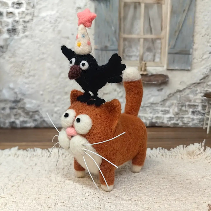 Side view of a handmade ginger cat wool felt figurine with an upright cream-tipped tail and a black crow wearing a star-topped party hat standing on its head.