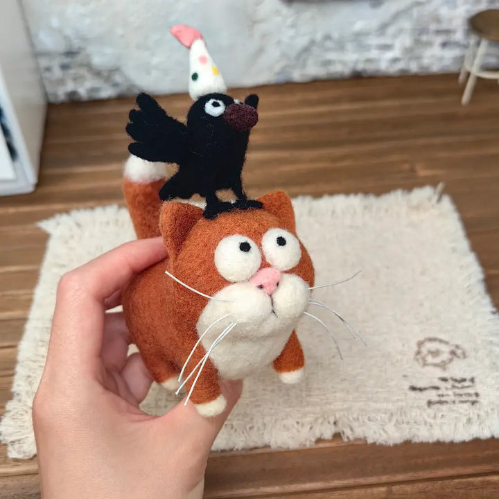 Hand holding a needle-felted ginger cat figurine with oversized eyes, white whiskers, and a black party crow perched on its head, showing the handmade details.
