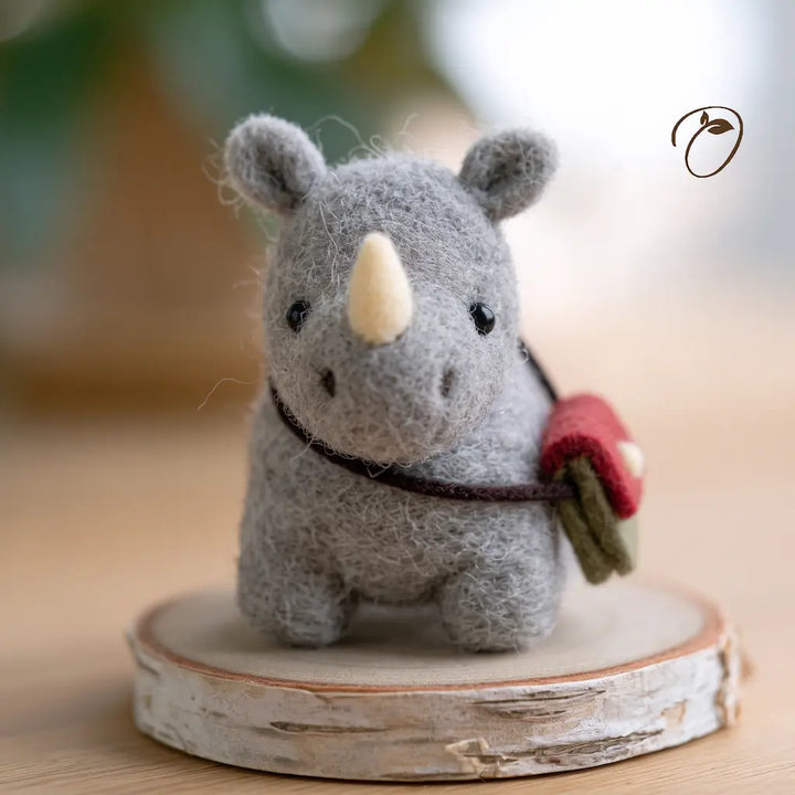 Front view of a gray wool felt rhino with a cream horn and red-and-green satchel, displayed on a round wood slice.