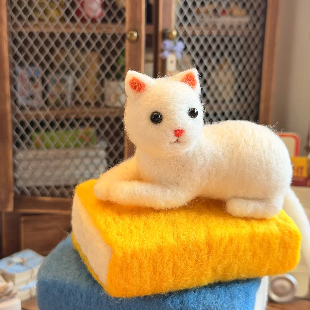 A white felted wool cat figurine with black eyes and a pink nose, lying on top of a yellow book-shaped cushion that sits on a blue block, set in front of a wooden wire-mesh cabinet.