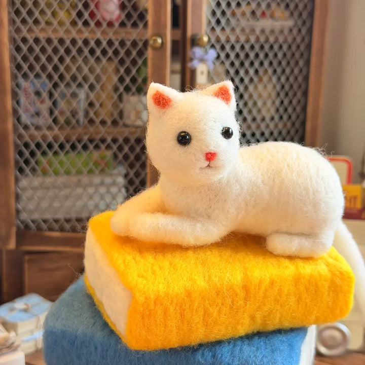 A white felted wool cat figurine with black eyes and a pink nose, lying on top of a yellow book-shaped cushion that sits on a blue block, set in front of a wooden wire-mesh cabinet.