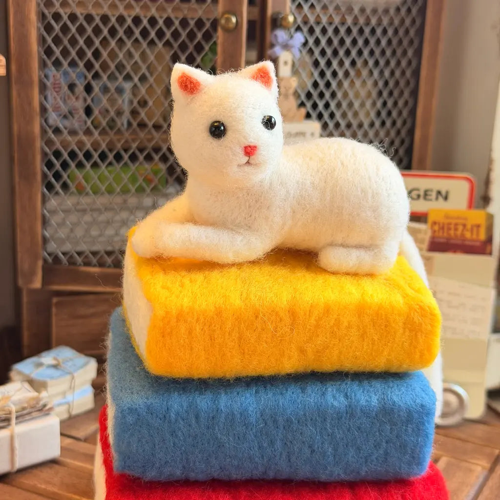 A white felted wool cat figurine with pink-tipped ears and black eyes, posed lying on top of a stack of three felted wool blocks in yellow, blue, and red, set against a warm, rustic background.