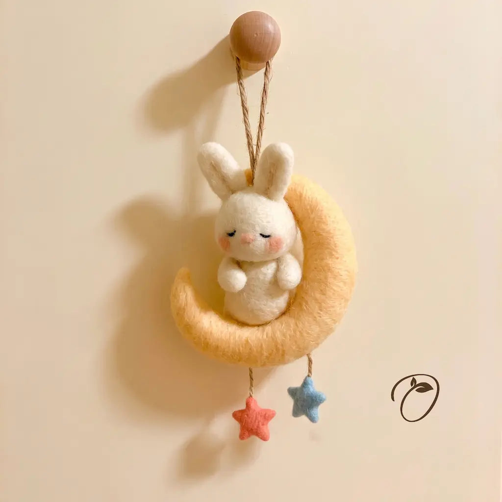 A handmade felt hanging ornament featuring a white bunny sitting on a crescent moon, with two dangling star charms in pink and blue, hanging from a wooden peg against a warm beige wall.