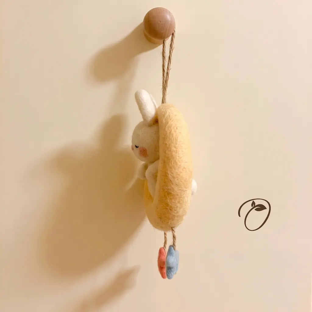 A small felted wool bunny hanging from a wooden peg on a beige wall. The bunny has white fur, a yellow body, and small dangling blue and pink accents.