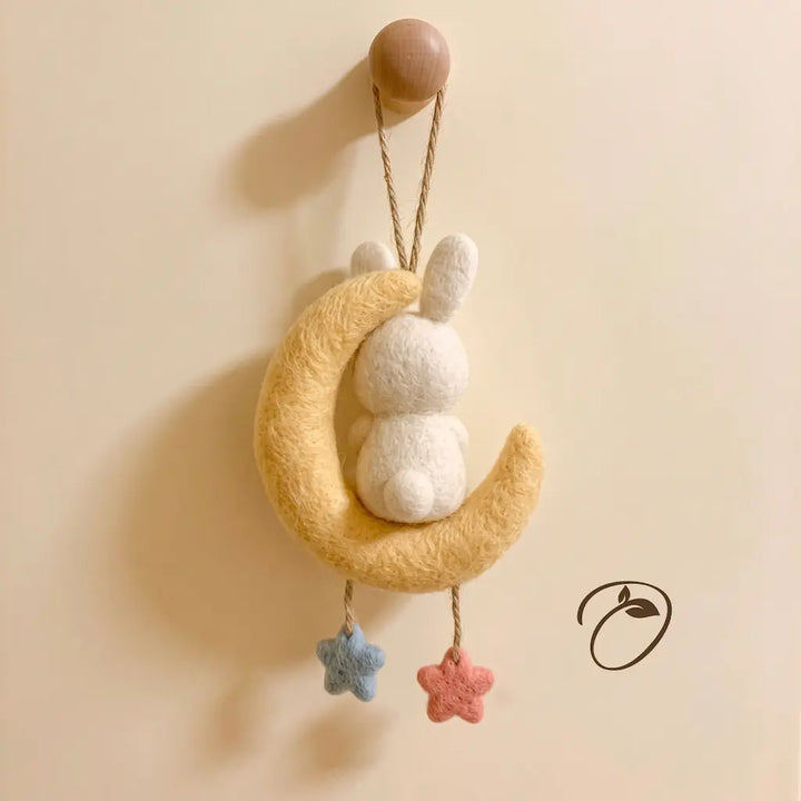 A handmade felt baby decoration featuring a white bunny sitting on a yellow crescent moon, hanging from a wooden peg on a beige wall. Two small star felt ornaments in blue and pink hang from the bottom.