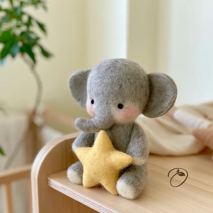 A small gray felt elephant toy holding a yellow felt star, sitting on a light wood surface in a softly blurred indoor setting.