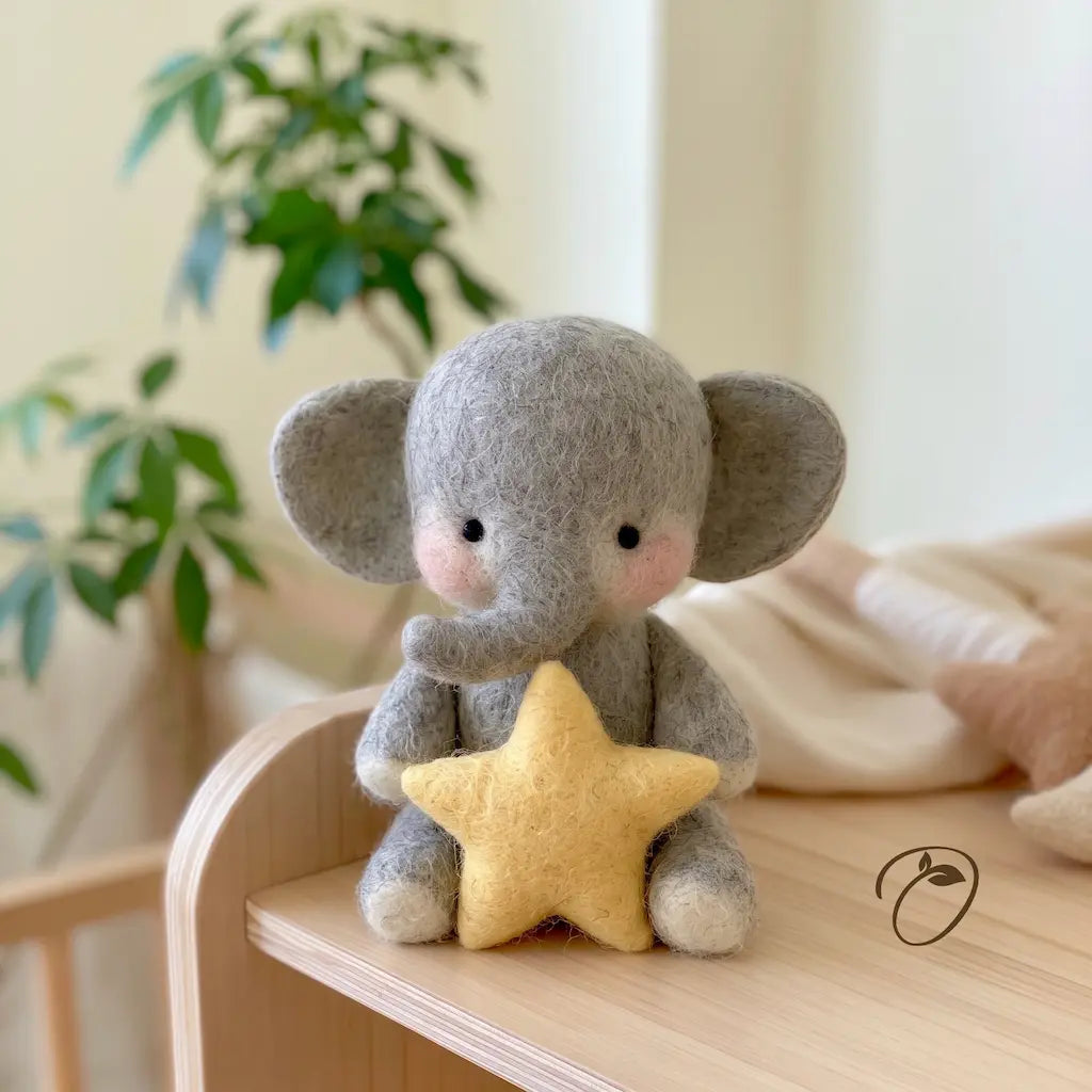 A small gray felt elephant toy holding a yellow felt star, placed on a light wood surface in a softly lit indoor setting with a blurred plant and neutral background.