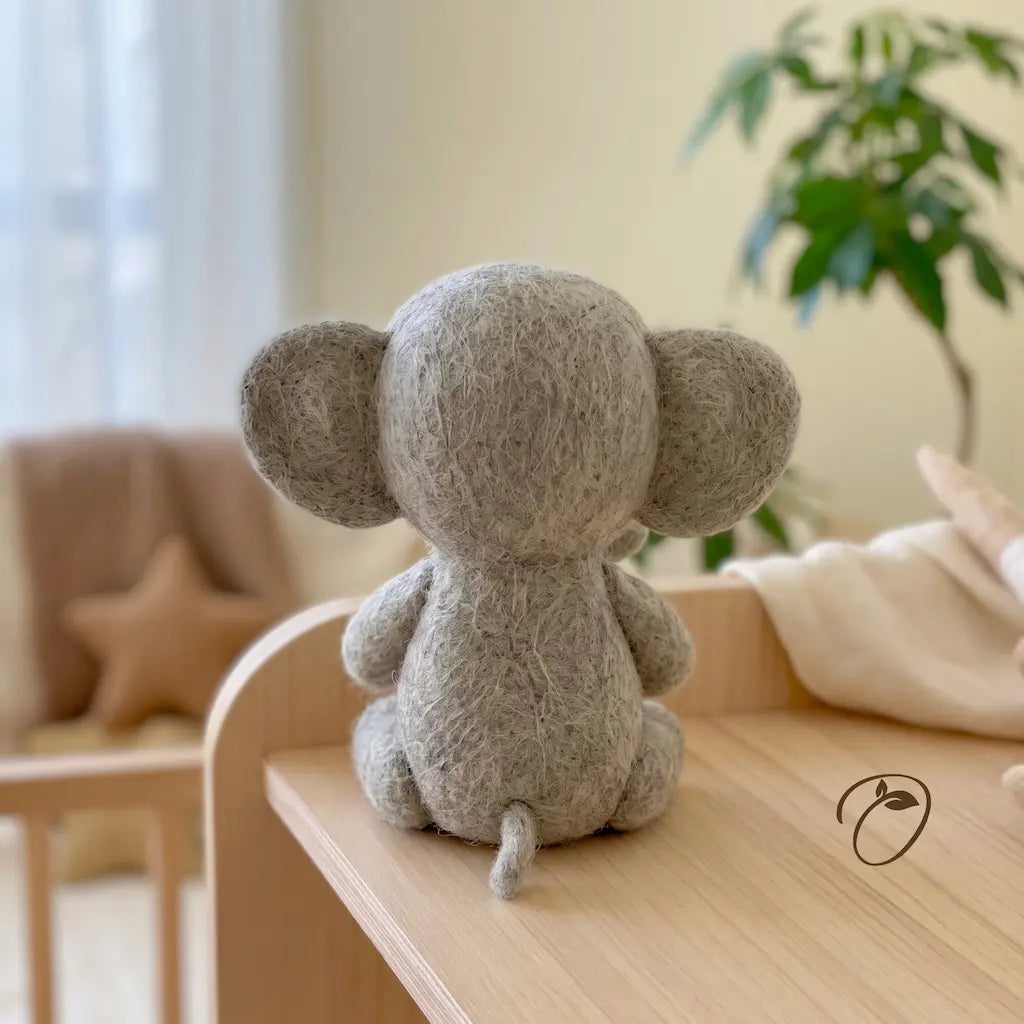 A gray felted wool mouse toy, shown from the back, sitting on a light wooden table in a softly lit nursery or playroom setting.