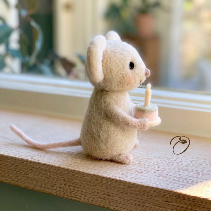 A small felted wool mouse figurine in cream-colored wool, holding a candle, sitting on a wooden windowsill with a softly blurred background.