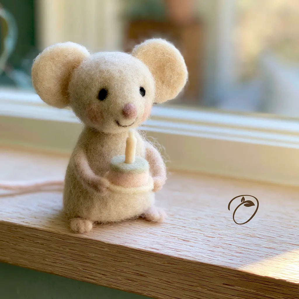 A small felted wool mouse figurine with large ears, black bead eyes, and a pink nose, sitting on a wooden windowsill and holding a tiny candle. The background shows a softly blurred window scene.
