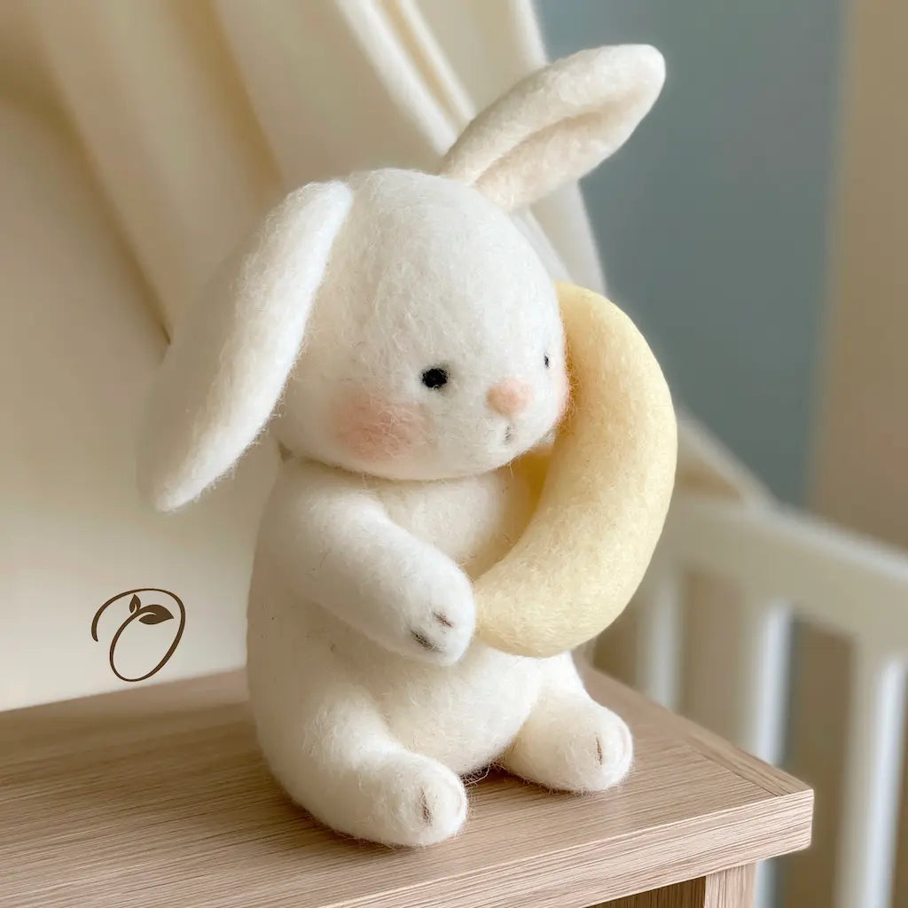 A small white felt rabbit plush toy sitting on a wooden table and holding a pale yellow crescent-shaped cushion, photographed in a softly lit indoor setting.