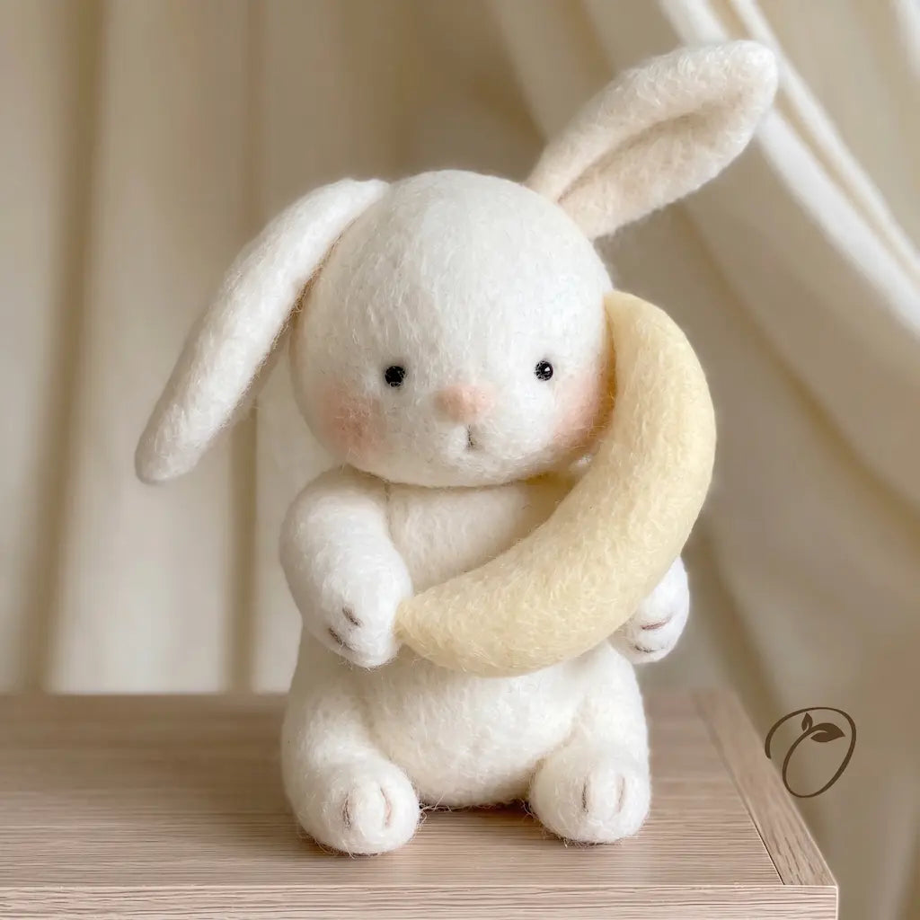 A small white felt rabbit toy holding a pale yellow crescent moon, sitting on a light wood surface with soft cream curtains in the background.