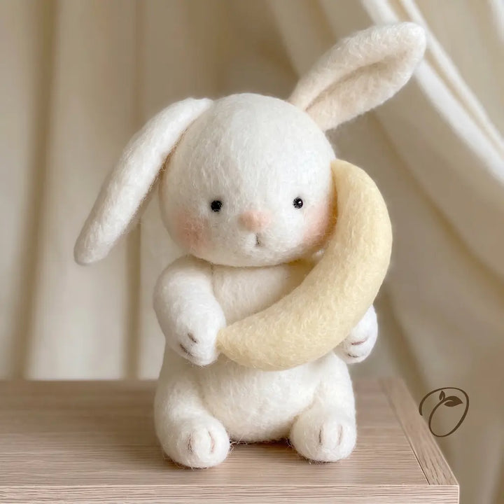 A small white felt rabbit toy holding a pale yellow crescent moon, sitting on a light wood surface with soft cream curtains in the background.
