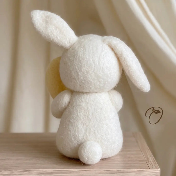A white felted wool bunny toy with long floppy ears, shown from the back on a light wood surface against a soft cream fabric backdrop.