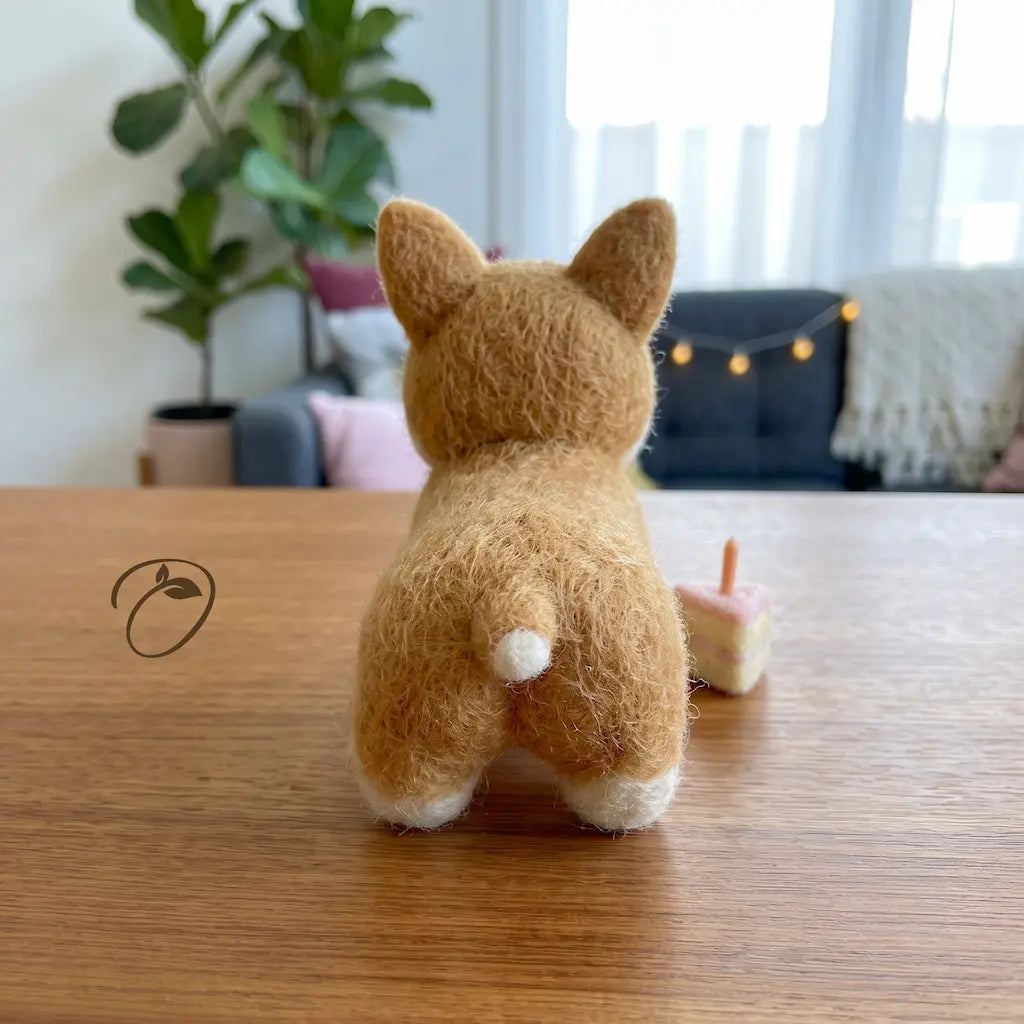 A small felt Corgi dog figurine, shown from behind on a wooden table in a cozy, softly lit room with a plant and string lights in the background.