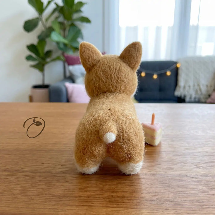 A small felt Corgi dog figurine, shown from behind on a wooden table in a cozy, softly lit room with a plant and string lights in the background.