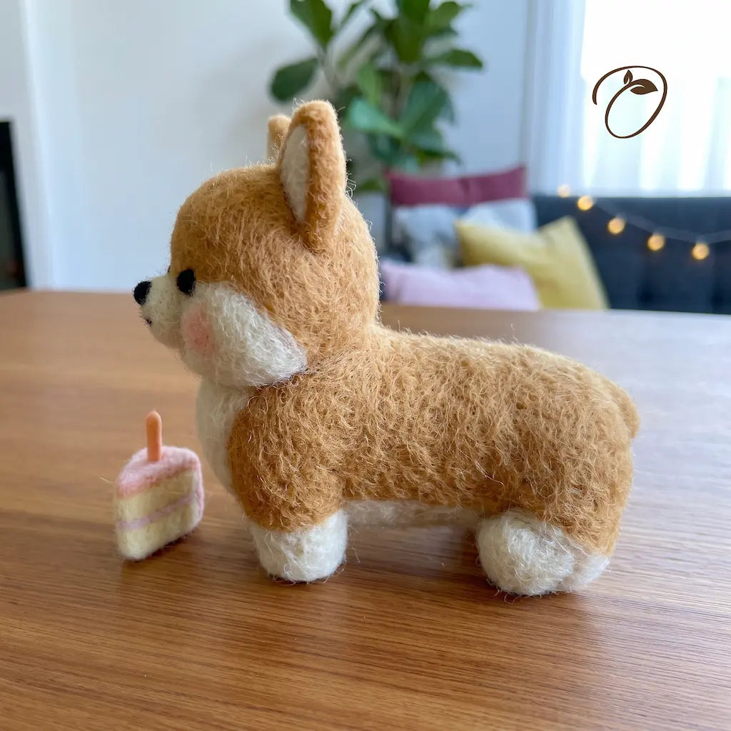 A small felt needle-art Shiba Inu figurine sits on a wooden table in a softly blurred indoor room. The toy is tan and cream with pink cheek details and a tiny pink-and-white cake piece beside it.