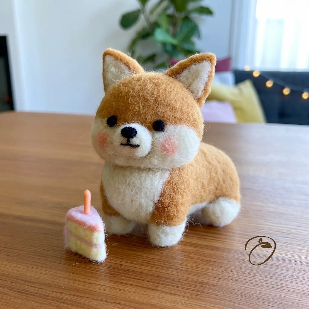 A small felt needle art dog figurine, resembling a Shiba Inu, sits on a wooden table next to a miniature pink and yellow cake. The background shows a softly blurred indoor room with a plant and warm string lights.