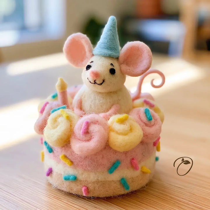 A cute felt mouse figurine wearing a light blue party hat, sitting on top of a pink and cream birthday cake decorated with pastel swirls and sprinkles, placed on a wooden tabletop.
