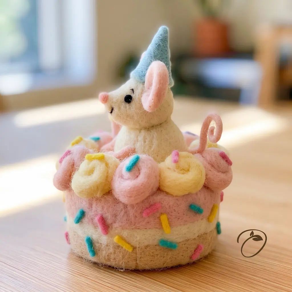 A cute felted wool mouse figurine wearing a light blue party hat, sitting on top of a pink and cream cupcake decorated with pastel donuts and colorful sprinkles, placed on a wooden tabletop.