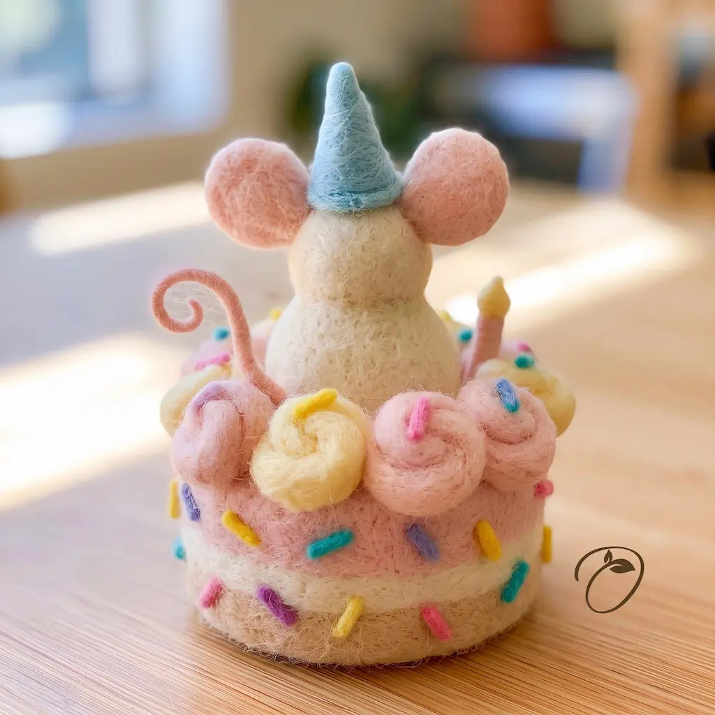 A cute handmade felt mouse figurine wearing a blue party hat and standing on a pink and cream tiered cake decorated with pastel swirls and colorful sprinkles, set on a wooden tabletop.