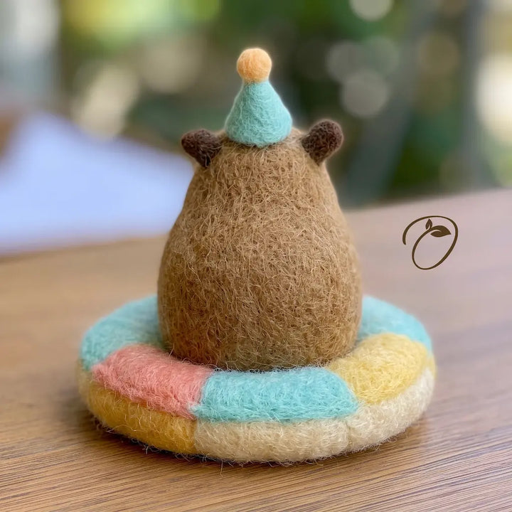 A small felted wool bear figurine sits on a round pastel cushion, photographed on a wooden surface with a softly blurred background. The bear is brown with a light blue party hat and the cushion is multicolored in pastel shades.