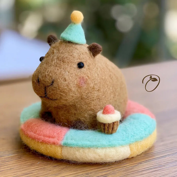 A cute felt capybara figurine sitting on a pastel rainbow donut float, holding a small cupcake. The capybara is made of brown wool felt and wears a mint green party hat with a yellow pom-pom.