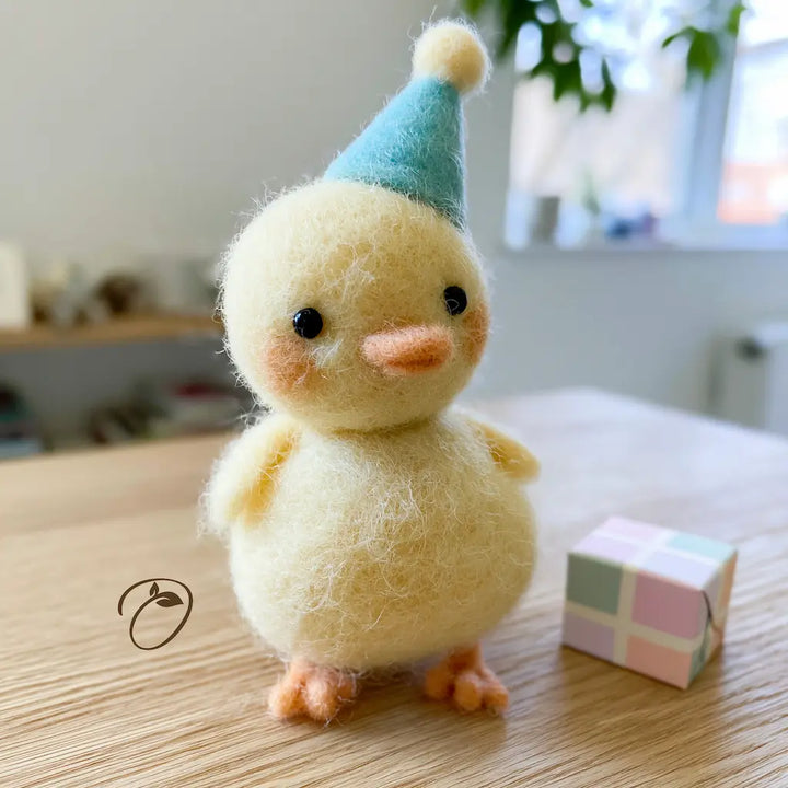 A cute needle-felted plush chick toy wearing a light blue party hat, standing on a light wooden table next to a small pastel checkered gift box.