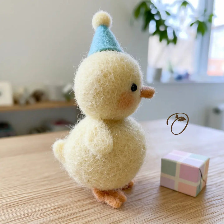 A fluffy needle-felted figurine duckling wearing a light blue party hat, standing on a light wood tabletop next to a small pastel gift box. The background is a softly blurred indoor room with a window and plant.