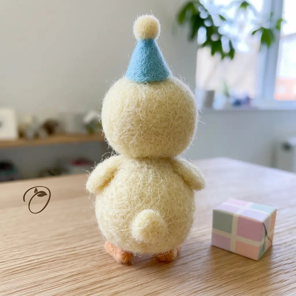 A handmade felt needle art duck figurine with a light blue party hat, standing on a wooden table next to a small pastel gift box.