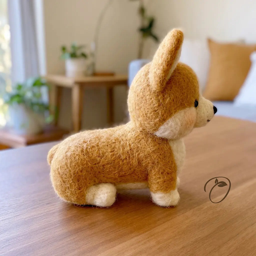 A small felted wool corgi dog figurine sits on a wooden table in a softly blurred indoor room. The toy has a warm tan body, cream paws and chest, black eyes, and a black nose.
