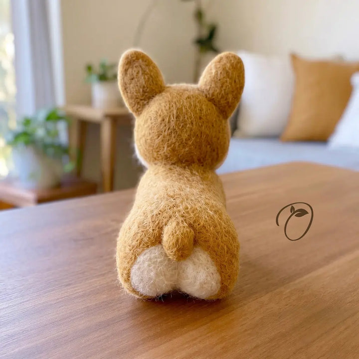 A small felt rabbit figurine viewed from behind, sitting on a wooden table in a softly blurred indoor setting with plants and cushions in the background.