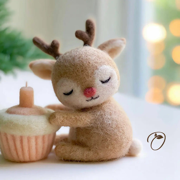 A cute needle-felted toy reindeer with brown antlers and a pink nose, sitting next to a small felted cupcake candle on a white surface with a soft, blurred holiday background.