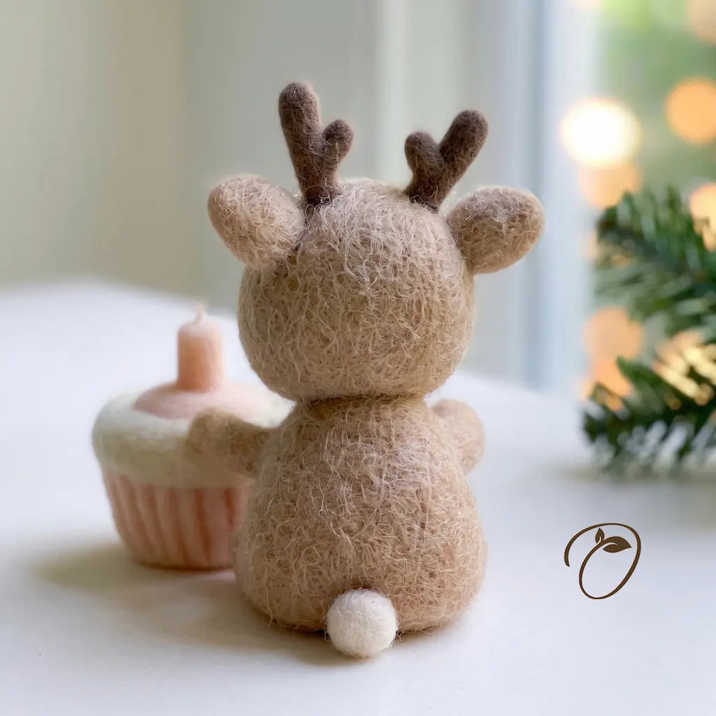 A small felted wool reindeer figurine shown from the back on a white surface, with a pink cupcake prop and soft holiday bokeh lights in the background.