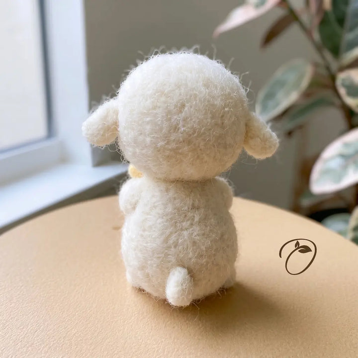 A small fluffy cream-colored felt sheep figurine shown from the back, sitting on a round tan surface near a window with a blurred plant in the background.