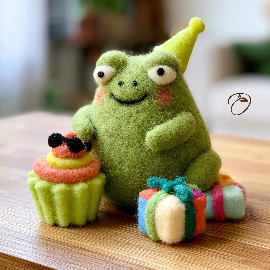 A green felt plush frog toy wearing a yellow party hat, sitting on a wooden surface with a small cupcake and wrapped gift beside it. The background is softly blurred and the image has a warm, cozy indoor feel.