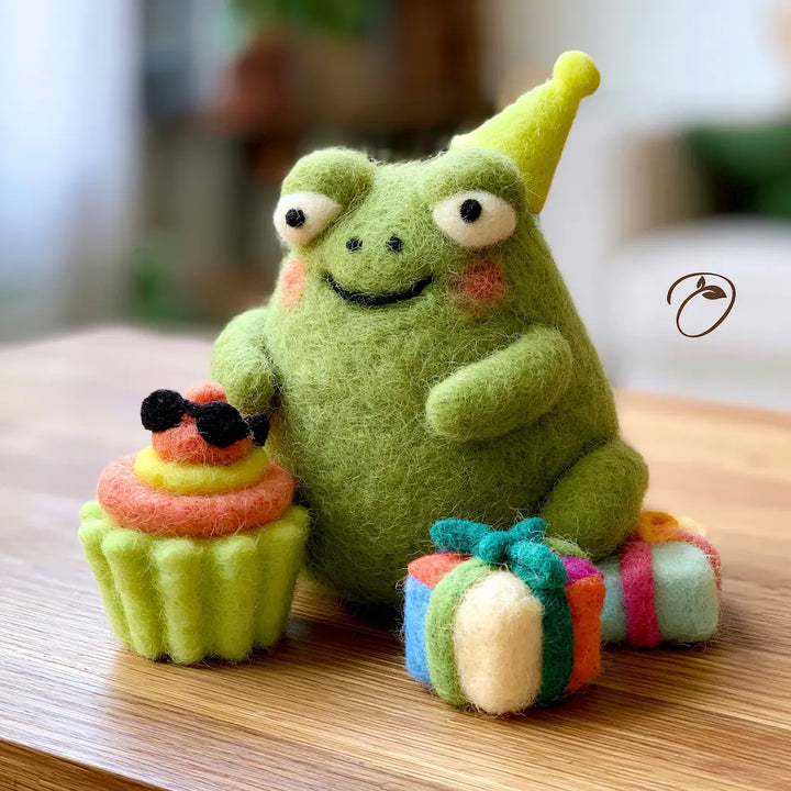 A green felt plush frog toy wearing a yellow party hat, sitting on a wooden surface with a small cupcake and wrapped gift beside it. The background is softly blurred and the image has a warm, cozy indoor feel.
